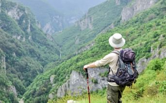 Hiker with backpack and walking stick overlooking the lush green Rakitnica gorge in Bosnia, surrounded by steep limestone cliffs and forest.