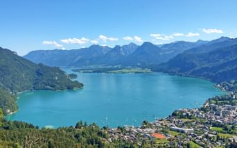 Aerial view of turquoise Wolfgangsee surrounded by mountains and green forests. A lakeside town is visible on the right shore under clear blue sky.