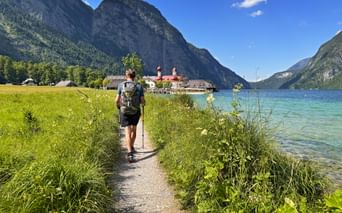 Hiker with backpack walking on path beside turquoise Königssee lake toward St. Bartholomä church with red onion domes, surrounded by Alps.