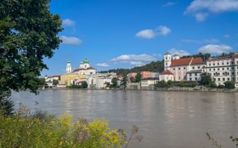 View of Passau's historic old town across the Danube River, showing baroque churches with green domes and colorful buildings under blue sky.