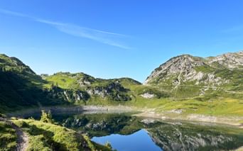 Bergsee Formarinsee spiegelt Felsgipfel unter blauem Himmel. Grüne Almwiesen und Wanderweg entlang des Ufers am Lechweg sichtbar.