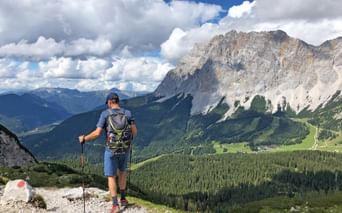 Wanderer mit Rucksack und Wanderstöcken auf felsigem Weg, mit Blick auf dramatisches Alpental mit Zugspitze-Gebirge und grünen Wäldern.