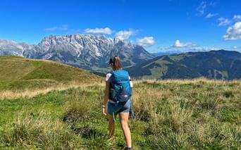 Female hiker with blue backpack walking on grassy alpine meadow with dramatic mountain peaks in background under blue sky with clouds.