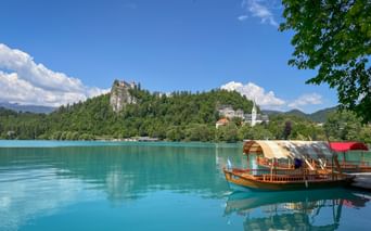 Traditional wooden boat on turquoise Lake Bled with Bled Castle on a forested cliff and a white church tower visible on the shore.