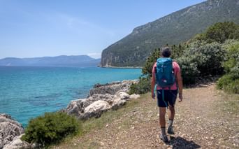 Hiker with backpack walking on rocky coastal path in Northern Dalmatia. Turquoise Adriatic Sea and green mountain slope visible.