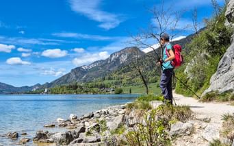 Female hiker with red backpack standing on rocky shore of Wolfgangsee lake in Salzkammergut, Austria, with mountains and blue sky in background.