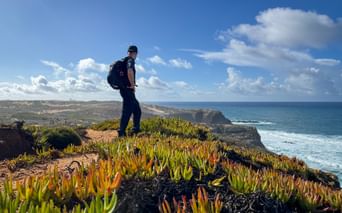 Wanderer mit Rucksack auf Klippe mit bunten Sukkulenten mit Blick auf den Atlantik an der Rota Vicentina in Portugal.