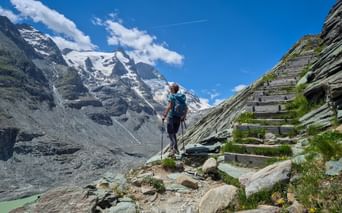 Wanderin mit Rucksack auf Bergweg an der Kaiser-Franz-Josefs-Höhe mit Großglockner-Gletschergipfel und Holzstufen unter blauem Himmel.