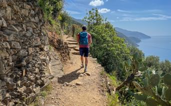 Hiker with blue backpack walking on stone path along Ligurian coast. Stone wall on left, Mediterranean Sea and green hillside on right.