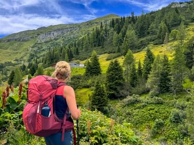 Frau mit rotem Rucksack wandert in grünem Alpental mit Nadelwäldern und Berghängen unter blauem Himmel.