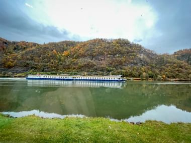 White and blue river cruise ship sailing on calm Danube waters with autumn-colored hills in background under cloudy sky.