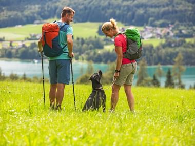 Zwei Wanderer mit Rucksäcken und einem schwarzen Hund auf einer grünen Wiese im Salzkammergut mit Blick auf einen türkisen See und Bergdorf.