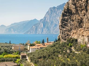 View of Torbole village at Lake Garda with church tower, olive groves, and dramatic rocky cliffs. Mountains rise across the blue lake.
