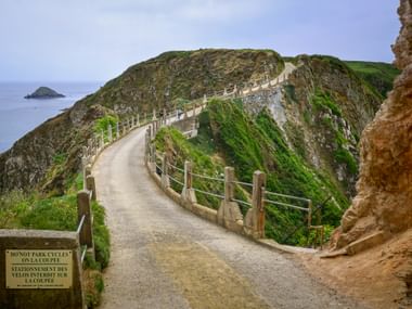 Gewundener Küstenweg auf Sark mit Holzgeländer entlang steiler grüner Klippen. Eine kleine Insel ist im blauen Meer sichtbar.