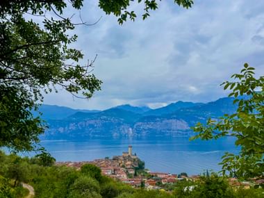 Blick auf Malcesine mit seiner mittelalterlichen Burg am Gardasee, eingerahmt von grünem Laub. Berge erheben sich im Hintergrund über dem blauen See.