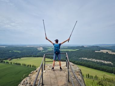 Hiker with backpack and trekking poles stands on Lilienstein summit platform with arms raised, overlooking green fields and forests below.