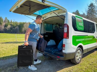 Man loading black suitcase into white Eurohike luggage transport van with green branding. Open rear doors show cargo space with backpacks.