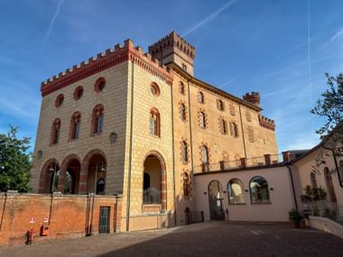 Barolo Castle courtyard in Piedmont with cream-colored facade, red brick accents, arched windows, and crenellated tower under blue sky.