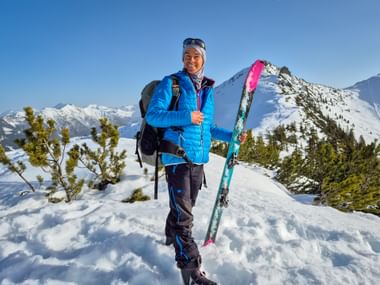 Frau in blauer Jacke mit Skiern auf verschneitem Berggipfel mit Panoramablick auf die Alpen und klarem blauen Himmel im Hintergrund.