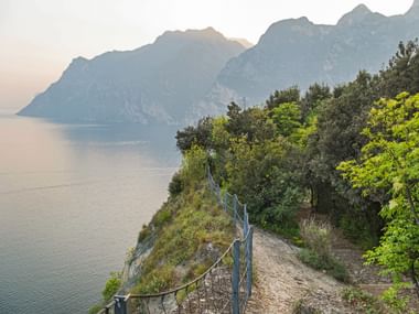 Narrow hiking path on Monte Brione with safety railing overlooking Lake Garda. Mountains rise in the background across calm waters.