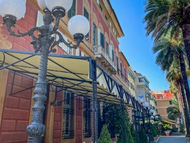 Street view in Sestri Levante with colorful Italian buildings, ornate street lamp, metal awning, and tall palm trees under blue sky.