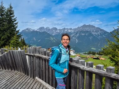 Lächelnde Wanderin in blauer Jacke auf hölzerner Aussichtsplattform mit Alpenpanorama und Tal mit traditionellen Gebäuden im Hintergrund.