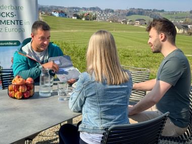 Eurohike staff member showing hiking map to couple at outdoor table with rolling hills and village in background. Banner displays eurohike.at branding.