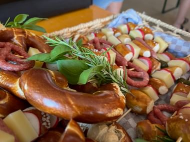 Bayerische Brotzeit mit Brezeln, Würsten, Käsescheiben und frischen Kräutern auf einem Weidenkorb bei einem Sommerfest.