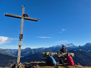 Hiker on wooden bench at Bodenberg summit cross with backpack and hiking poles, overlooking Dachstein mountain range under blue sky.