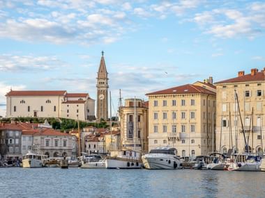 Coastal view of Piran, Slovenia showing historic bell tower, Mediterranean architecture, and marina with sailboats and yachts under cloudy sky.
