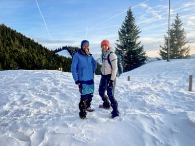 Zwei Frauen in Winterkleidung mit Schneeschuhen auf verschneitem Weg auf der Schafbergalm. Bergwald und blauer Himmel mit Kondensstreifen im Hintergrund.