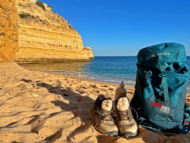 Hiking boots and backpack on sandy beach at Praia da Marinha, Algarve. Golden cliffs rise from turquoise water under blue sky.