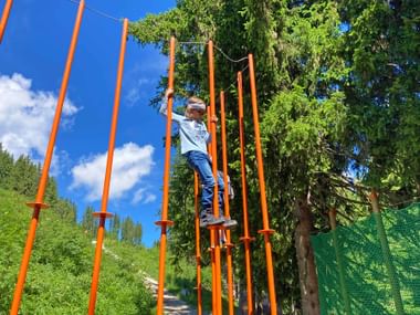 Child climbing orange poles at Schmidolin motor skills playground on Schmittenhöhe near Zell am See, surrounded by green trees and blue sky.