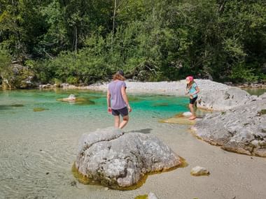 Zwei Wanderer waten im kristallklaren türkisfarbenen Soča-Fluss in den Julischen Alpen. Große weiße Felsen im flachen Wasser, Kiesstrand und Wald.