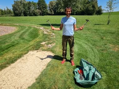 Lächelnde Person im blauen Shirt hält Gartengeräte in beiden Händen, steht auf Grasweg mit grüner Tasche unter blauem Himmel.