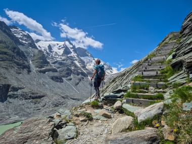 Hiker with backpack on rocky mountain trail at Kaiser Franz Josefs Höhe with snow-capped glacier peaks and blue sky in background.