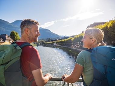 Two hikers with backpacks standing on a bridge in Bad Ischl, looking at each other. River and mountains visible in the background.