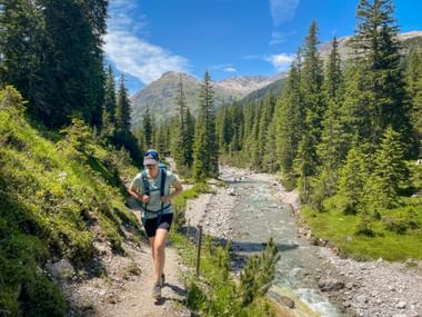Wanderin mit Rucksack und Wanderstöcken am Lechweg neben türkisfarbenem Gebirgsfluss, umgeben von Nadelwald und Alpengipfeln.