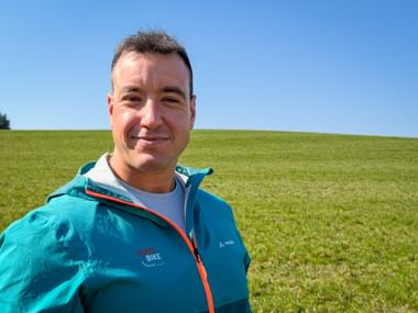 Mann in türkisfarbener Eurobike-Jacke lächelt in die Kamera mit grüner Wiese und blauem Himmel im Hintergrund.