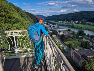 Hiker with blue backpack on viewing platform of Bad Schandau historic elevator, overlooking Elbe River valley with town and forested hills.
