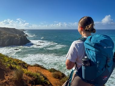 Wanderin mit blauem Rucksack betrachtet felsige Klippen und Atlantik bei Praia dos Carriços nahe Almograve an der Rota Vicentina im Alentejo.