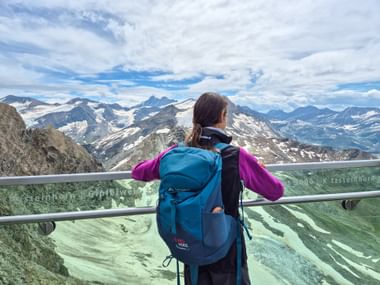 Wanderin mit blauem Rucksack auf der Aussichtsplattform am Kitzsteinhorn mit Blick auf schneebedeckte Gipfel der Hohen Tauern.