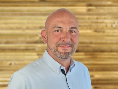 Portrait of Ricardo Gouveia with a bald head and gray beard, wearing a light blue shirt, smiling in front of a wooden wall.