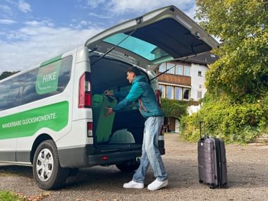 Eurohike Gepäcktransfer-Service-Van Mann lädt Gepäck in weißen und grünen Eurohike-Van mit offenen Hecktüren. Schwarzer Koffer steht auf Gehweg mit Haus im Hintergrund.