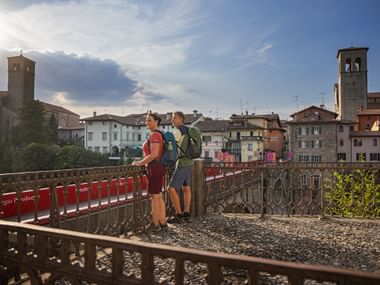 Two hikers with backpacks standing on ornate metal bridge in Cividale del Friuli, with historic buildings and bell towers in background.