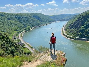 Wanderer mit rotem Rucksack auf Felsvorsprung mit Blick auf das Rheintal mit bewaldeten Hügeln und Frachtschiffen auf dem Wasser.