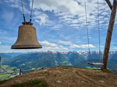 Bronzene Wunschglocke und Holzschaukel am Rosskopf mit Panoramablick auf schneebedeckte Alpengipfel und grüne Täler.