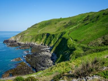 Gewundener Küstenweg auf grünen Klippen über felsiger Küste mit blauem Meer. Der Pfad führt entlang steiler Hänge zwischen Bonne Nuit Bay und Bouley Bay.