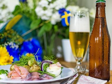 Teller mit Surströmming, Kartoffeln, Zwiebeln und Kräutern neben Bierflasche und Glas, mit schwedischer Flagge und Blumen im Hintergrund.