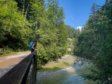Female hiker with blue backpack standing on bridge railing, overlooking Zinkenbach river in Salzkammergut. Dense forest and blue sky visible.
